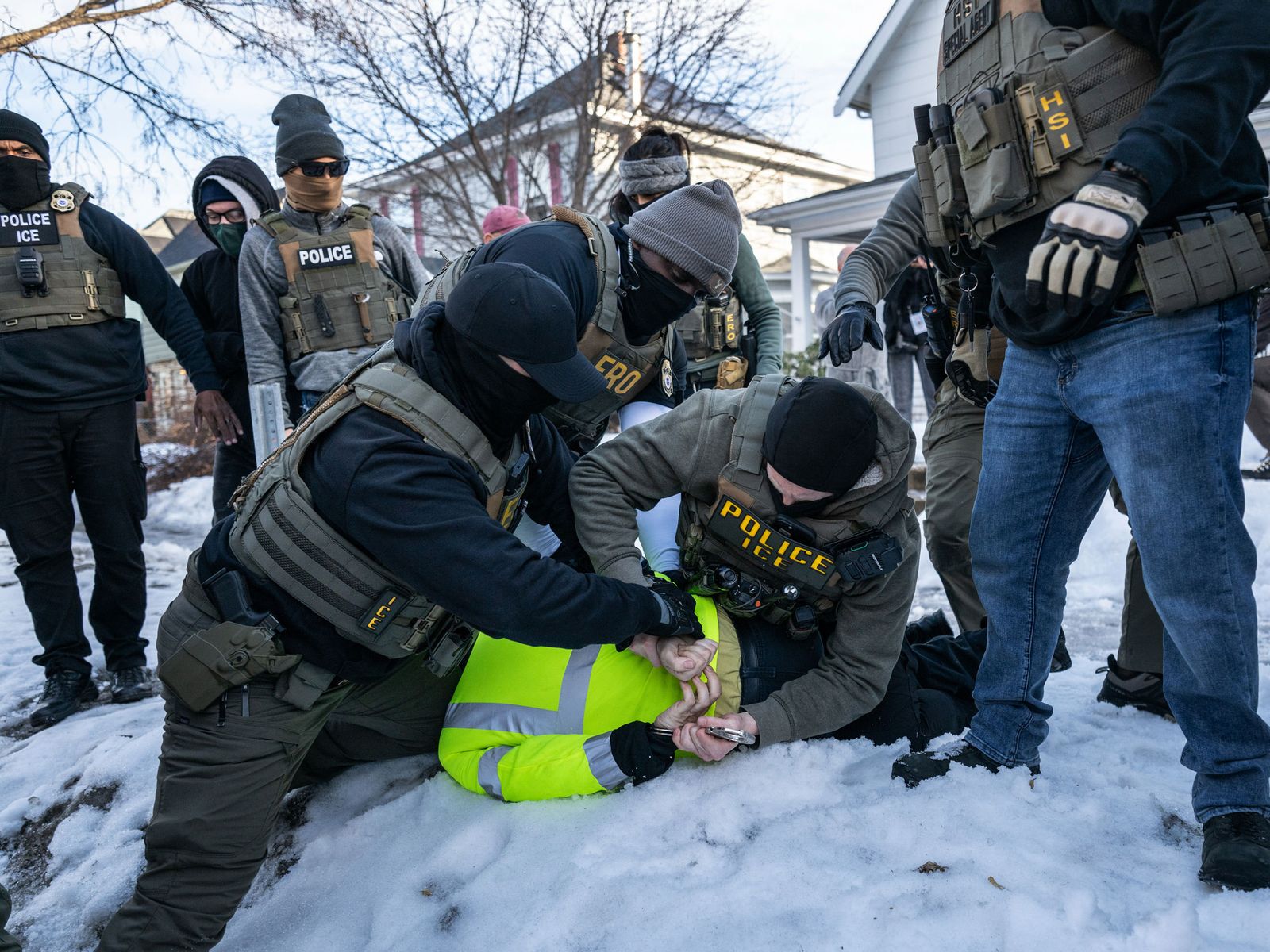 Several ICE agents restraining a person wearing yellow vest to the ground on snow.