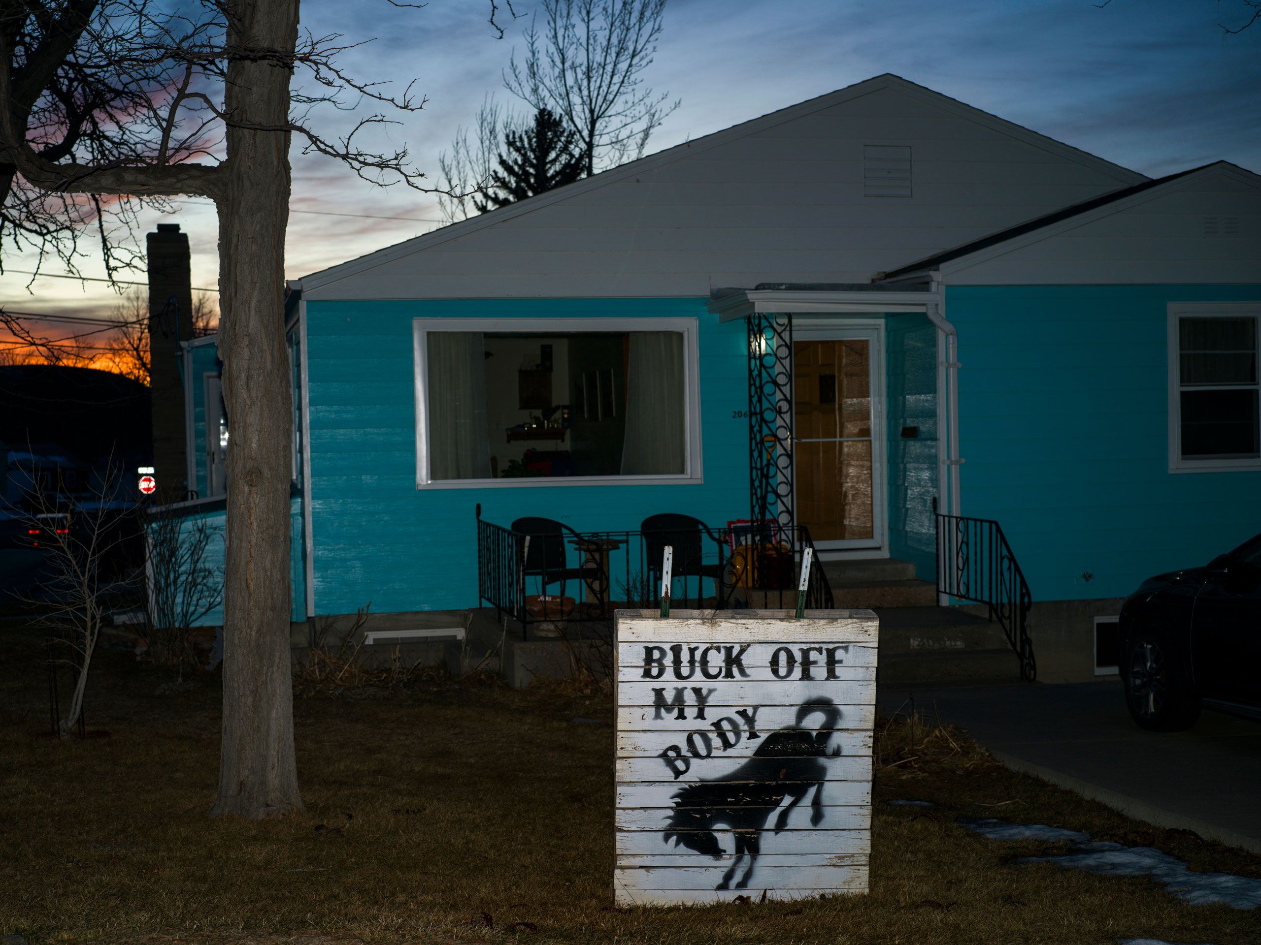 A blue house with a white wooden sign on a lawn which features a horse and the words “Buck off my body.”